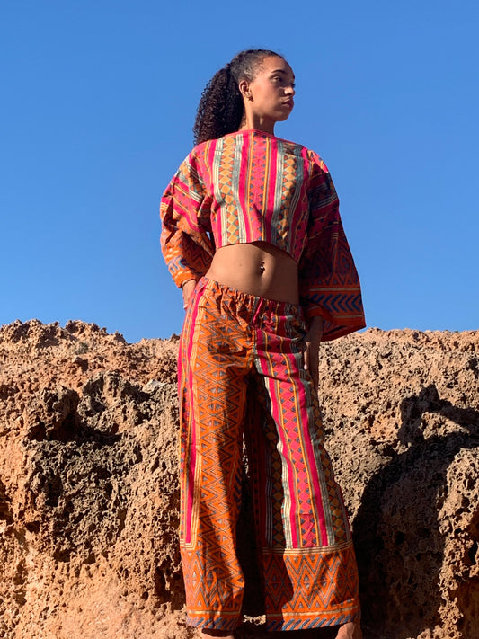 Model in pink and orange striped upcycled cotton two-piece with metallic gold print, featuring oversized crop top and wide-leg trousers, shot on sunlit coastal rocks.