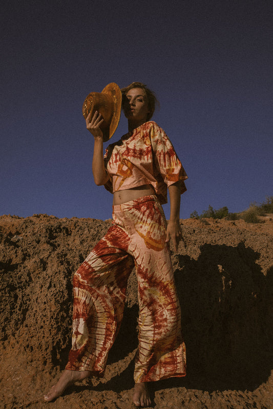 Model wearing an orange and cream tie-dye African batik two-piece set with oversized crop top and wide-leg trousers, standing barefoot on rugged Ibiza coastline.