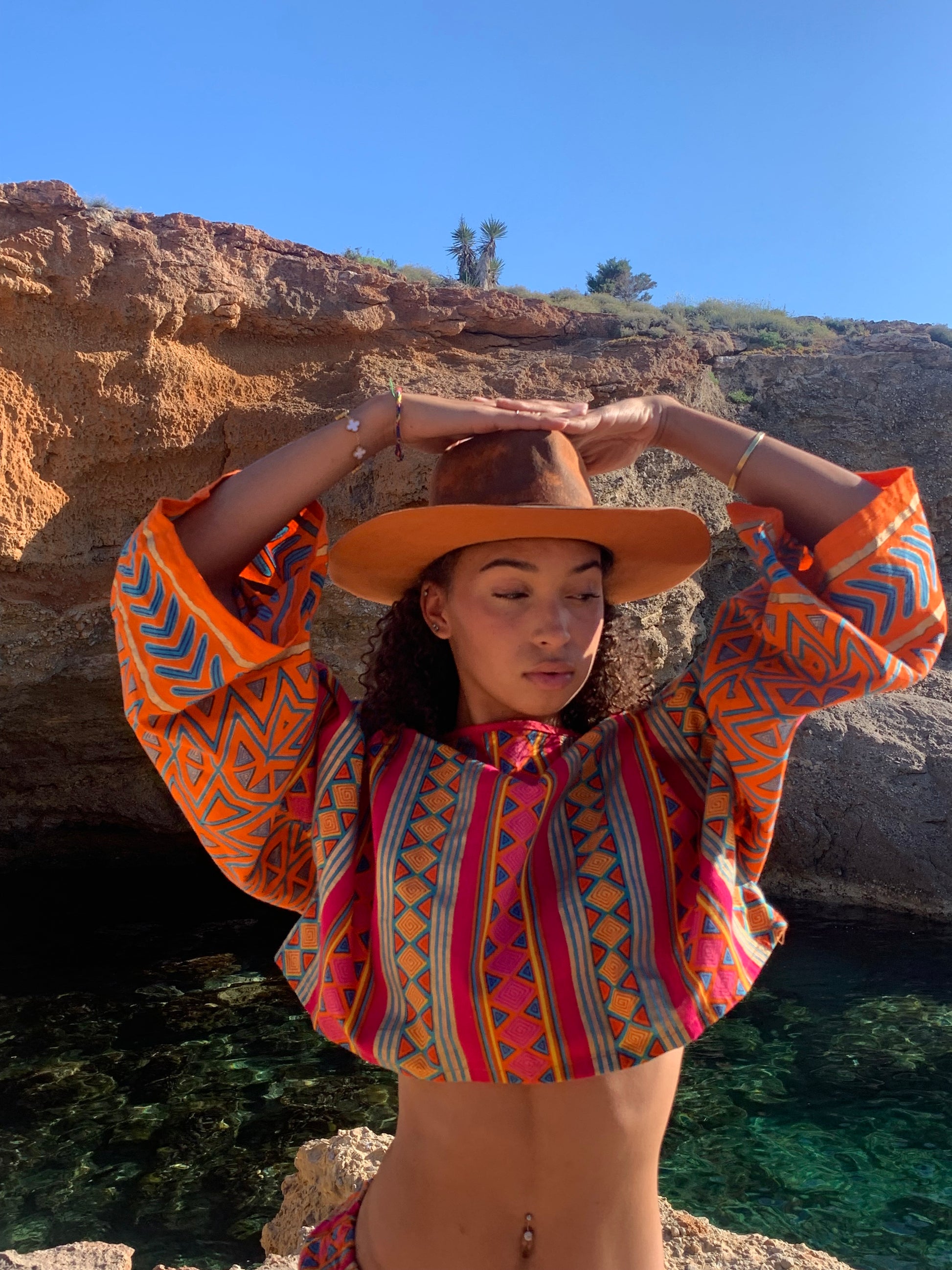 Model in pink and orange striped upcycled cotton two-piece with metallic gold print, featuring oversized crop top and wide-leg trousers, shot on sunlit coastal rocks.Model in pink and orange striped upcycled cotton two-piece with metallic gold print, featuring oversized crop top and wide-leg trousers, shot on sunlit coastal rocks.