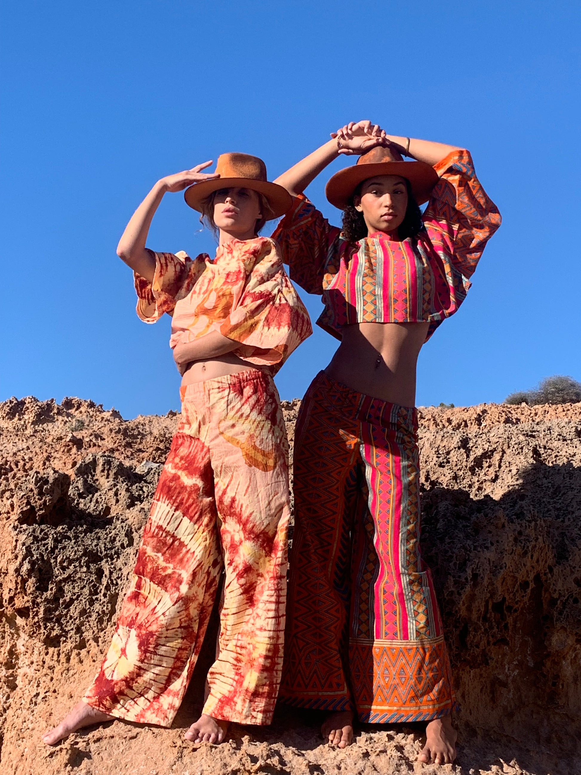 Model in pink and orange striped upcycled cotton two-piece with metallic gold print, featuring oversized crop top and wide-leg trousers, shot on sunlit coastal rocks.