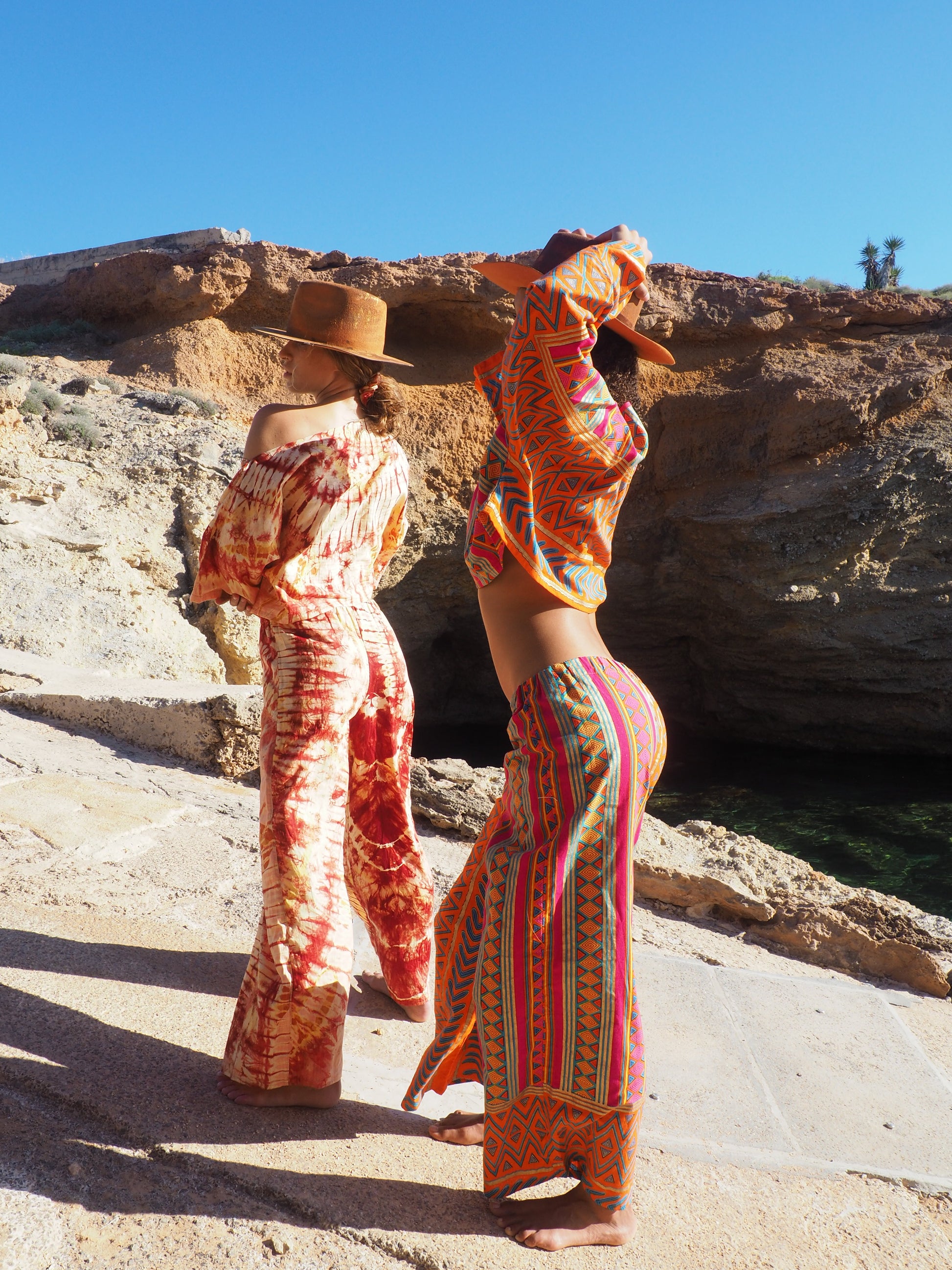 Model in pink and orange striped upcycled cotton two-piece with metallic gold print, featuring oversized crop top and wide-leg trousers, shot on sunlit coastal rocks.